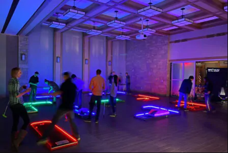 A group of people playing minigolf indoors on portable LED lit putt putt courses.